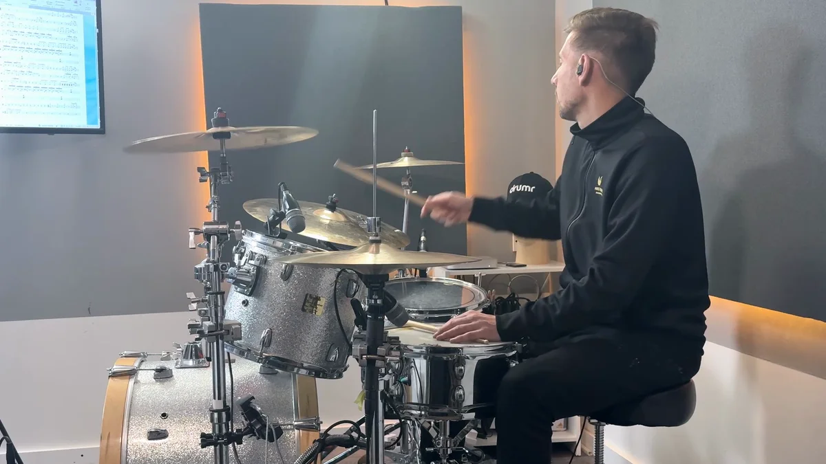 Geoff Fry in a studio swinging a stick into the cymbals of a silver-sparkle drum kit, with a Drumr cap and drum notation on a screen in the background