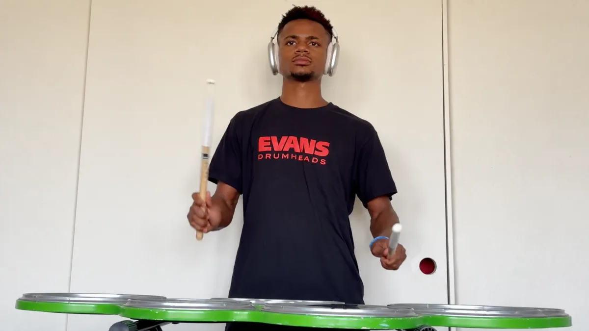 A tenor drummer wearing headphones, practicing on a set of marching tenors against a white wall